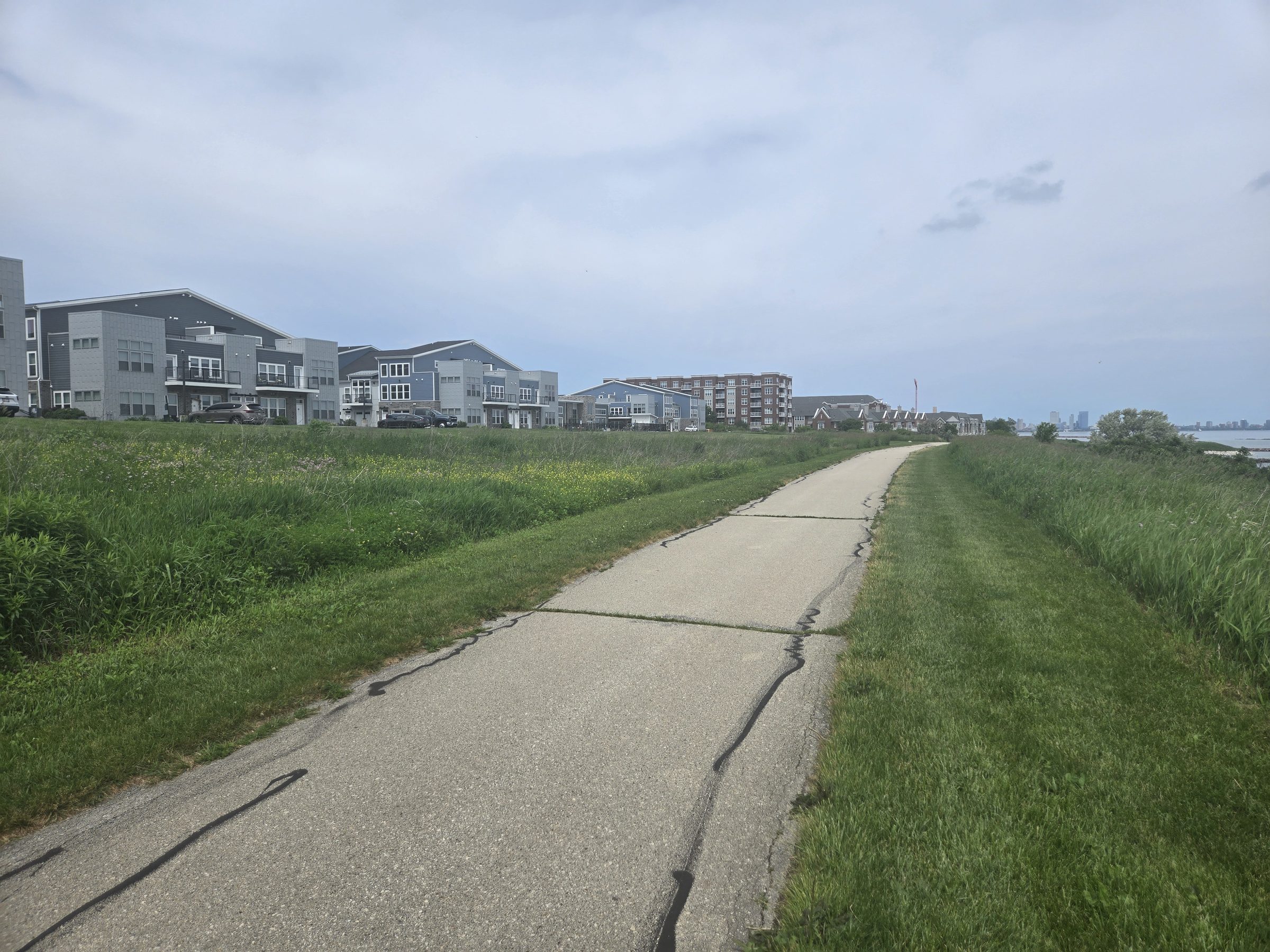 The lakefront walking path in St. Francis: a paved trail along the grassy bluff above Lake Michigan, homes on the left and the Milwaukee skyline faint in the distance.